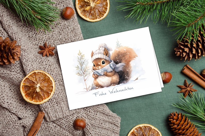 K&ouml;nnte beinhalten: Eine wei&szlig;e Karte mit einer Aquarell-Illustration eines Eichh&ouml;rnchens, das einen wei&szlig;en Strickhut und einen blauen Schal tr&auml;gt. Das Eichh&ouml;rnchen steht im Schnee mit einem Kiefernbaum im Hintergrund. Die Karte sagt "Frohe Weihnachten".