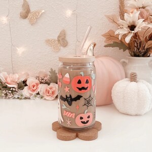 K&ouml;nnte beinhalten: Ein durchsichtiger Glasbecher mit Bambusdeckel und Strohhalm, verziert mit Halloween-Aufklebern. Die Aufkleber zeigen K&uuml;rbisse, eine Fledermaus, einen Geist und S&uuml;&szlig;igkeiten. Der Becher steht auf einem Untersetzer aus Kork.