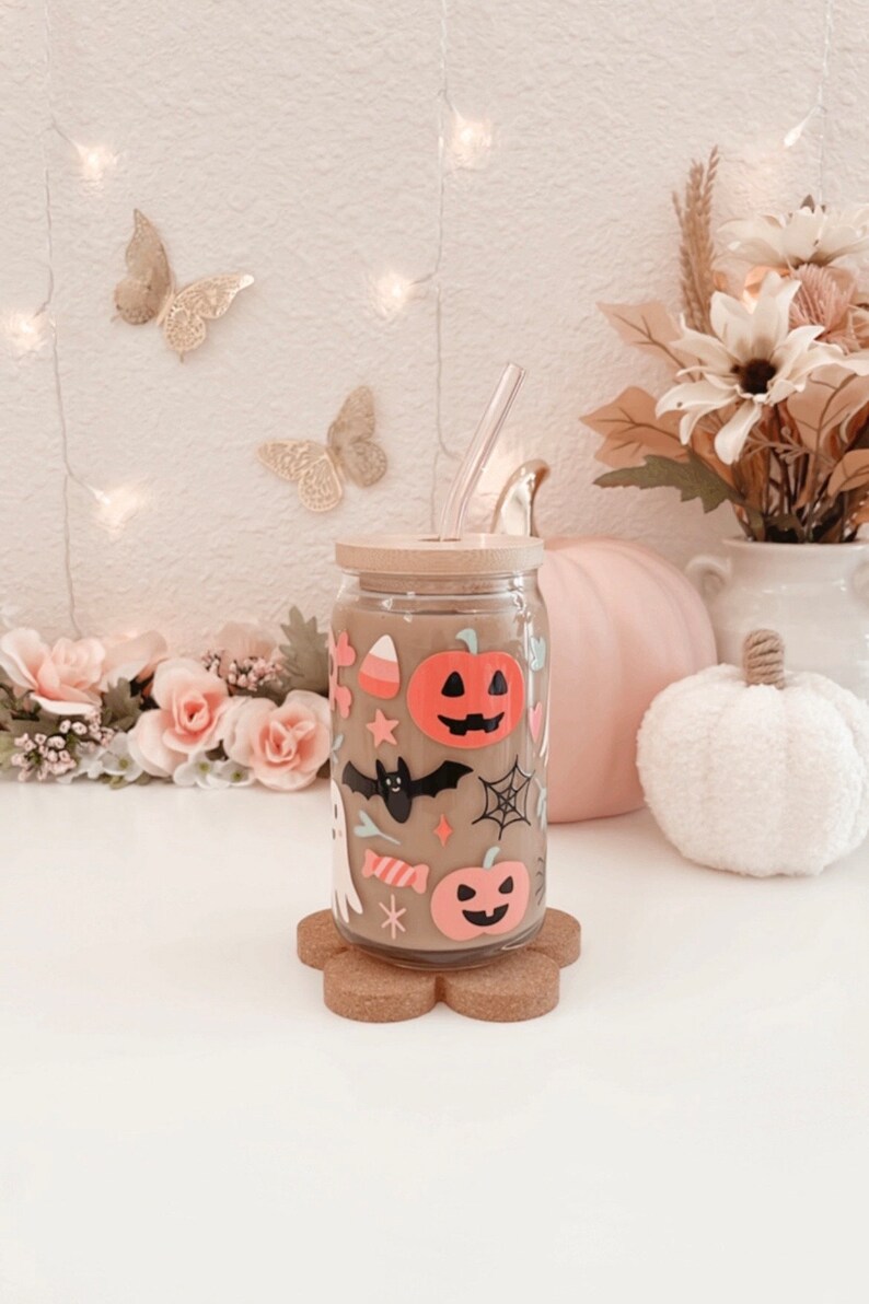 K&ouml;nnte beinhalten: Ein durchsichtiger Glasbecher mit Bambusdeckel und Strohhalm, verziert mit Halloween-Aufklebern. Die Aufkleber zeigen K&uuml;rbisse, eine Fledermaus, einen Geist und S&uuml;&szlig;igkeiten. Der Becher steht auf einem Untersetzer aus Kork.