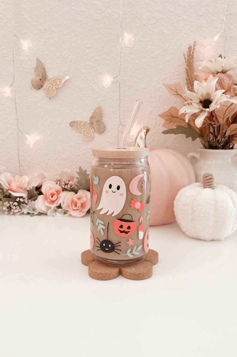 K&ouml;nnte beinhalten: Ein durchsichtiger Glasbecher mit Bambusdeckel und Strohhalm, verziert mit Halloween-Stickern, darunter ein Geist, eine Spinne und ein K&uuml;rbislaterne. Der Becher steht auf einem bl&uuml;tenf&ouml;rmigen Untersetzer aus Kork.