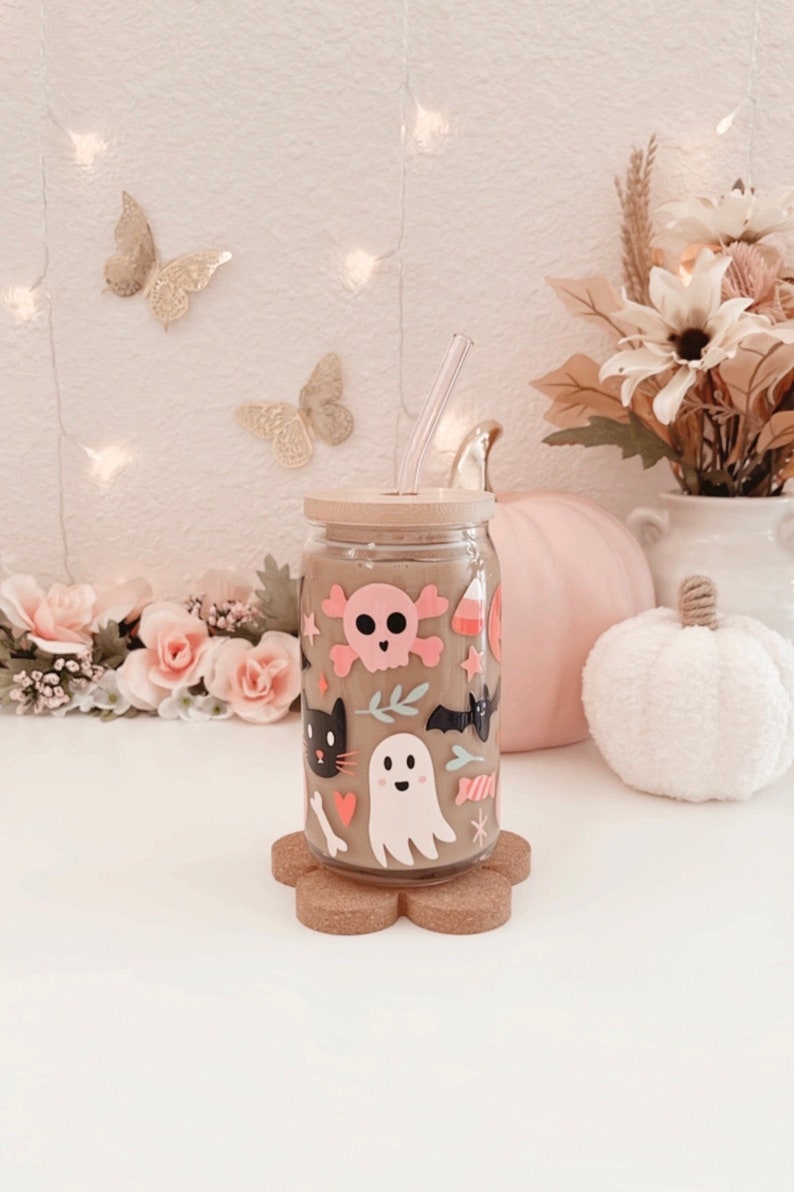 K&ouml;nnte beinhalten: Ein durchsichtiger Glasbecher mit Bambusdeckel und Strohhalm, verziert mit Halloween-Aufklebern. Die Aufkleber zeigen einen rosa Totenkopf, eine schwarze Katze, einen Geist und S&uuml;&szlig;igkeiten. Der Becher steht auf einem Untersetzer aus Kork.