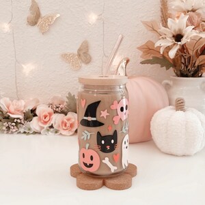 K&ouml;nnte beinhalten: Ein klares Glas mit Bambusdeckel und Strohhalm, verziert mit Halloween-Aufklebern. Die Aufkleber zeigen einen Hexenhut, eine schwarze Katze, einen K&uuml;rbis, einen Totenkopf und einen Geist. Das Glas steht auf einem blumenf&ouml;rmigen Korkuntersetzer.