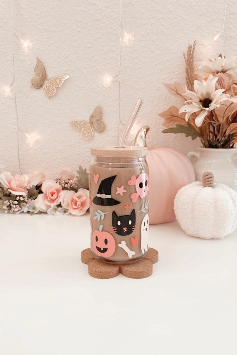 K&ouml;nnte beinhalten: Ein klares Glas mit Bambusdeckel und Strohhalm, verziert mit Halloween-Aufklebern. Die Aufkleber zeigen einen Hexenhut, eine schwarze Katze, einen K&uuml;rbis, einen Totenkopf und einen Geist. Das Glas steht auf einem blumenf&ouml;rmigen Korkuntersetzer.