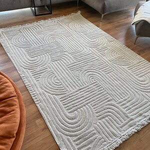 May include: A cream-colored rug with a geometric pattern of raised lines and arches. The rug has a fringe border.