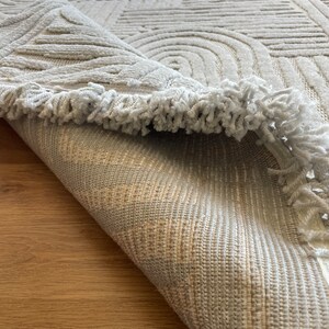 May include: A white rug with a geometric pattern and a fringe border. The rug is rolled up and the underside is visible.