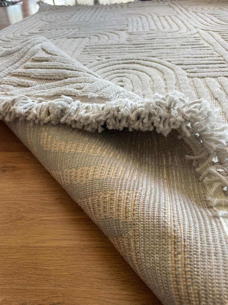 May include: A white rug with a geometric pattern and a fringe border. The rug is rolled up and the underside is visible.
