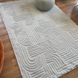 May include: A light gray area rug with a geometric pattern of curved lines and a fringe border. The rug is on a hardwood floor and a gray ottoman is sitting on top of the rug.