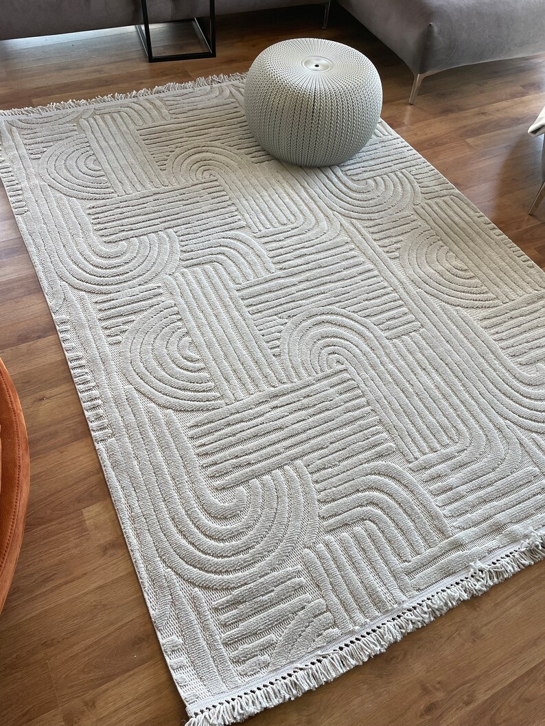 May include: A light gray area rug with a geometric pattern of curved lines and a fringe border. The rug is on a hardwood floor and a gray ottoman is sitting on top of the rug.