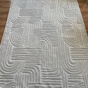 May include: A white area rug with a geometric pattern of raised lines and arches. The rug has a fringe border.