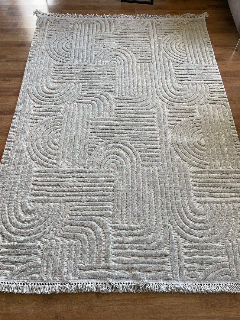 May include: A white area rug with a geometric pattern of raised lines and arches. The rug has a fringe border.