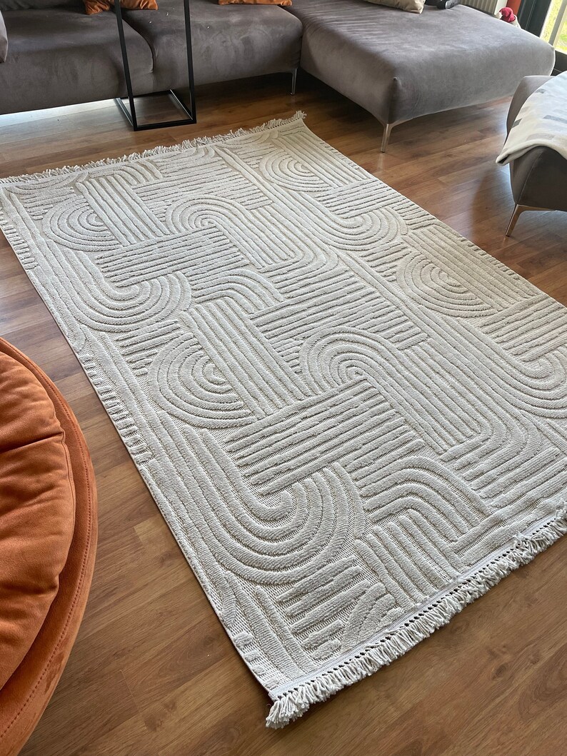 May include: A cream-colored rug with a geometric pattern of raised lines and arches. The rug has a fringe border.