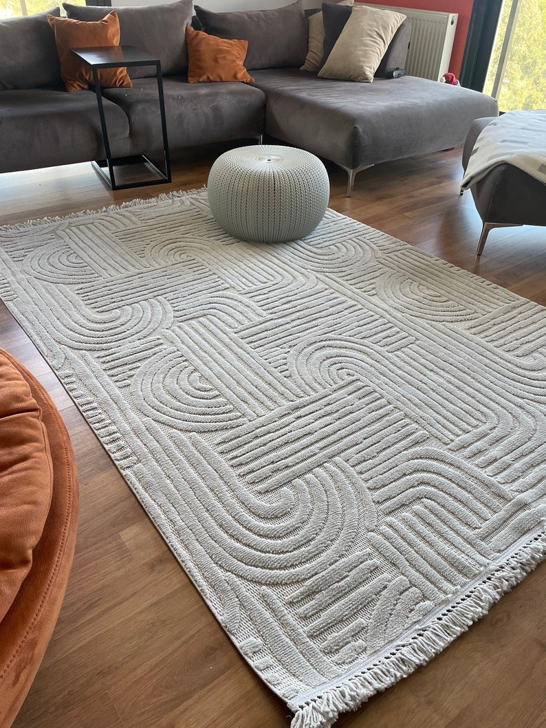 May include: A white, geometric patterned rug with a fringe border. The rug is laid out on a hardwood floor in a living room setting with a gray sectional sofa and a white ottoman.