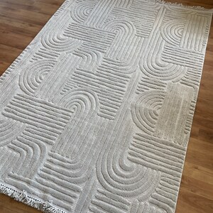 May include: A cream-colored rug with a geometric pattern of raised lines and arches. The rug has a fringe border.