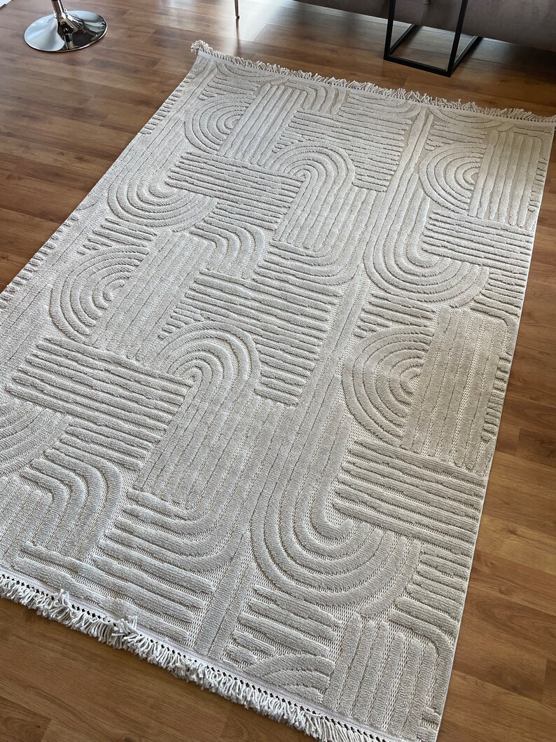 May include: A cream-colored rug with a geometric pattern of raised lines and arches. The rug has a fringe border.