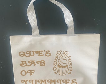 Könnte beinhalten: Weiße Canvas-Tragetasche mit goldener Glitzerbeschriftung "Que's Bag of Yummmies". Rechts neben dem Text befindet sich eine goldene Glitzer-Cupcake-Grafik.