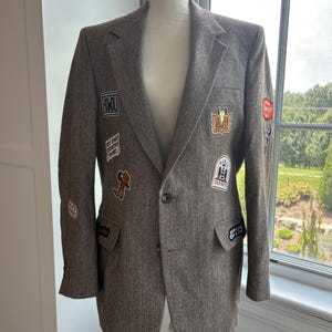 May include: A brown herringbone blazer featuring a single-button closure, notched lapels, and multiple embroidered patches. The patches include text such as "Saloon Western," "But First Country Music," and "Cowboys," along with images of a boot and skull.