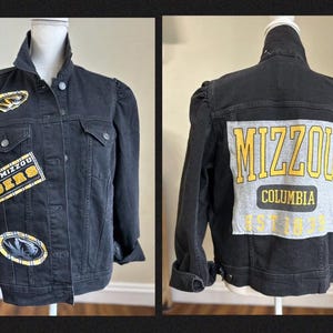 May include: A black denim jacket with puff sleeves, featuring Mizzou Tigers patches on the front and a large grey panel on the back with the yellow text MIZZOU COLUMBIA EST 1839. The jacket has a button-up front, collar, and chest pockets.