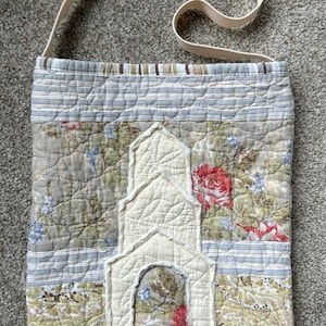 May include: A quilted fabric shoulder bag featuring a patchwork design of floral and striped patterns in muted tones of green, blue, and red. The bag has a cream-colored fabric applique shaped like a building with an arched doorway.