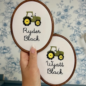 Puede incluir: Dos bastidores de bordado ovalados de madera con tela blanca y tractores verdes y amarillos bordados. El bastidor superior muestra el nombre Ryder Black en hilo negro, y el inferior muestra el nombre Wyatt Black en hilo negro.