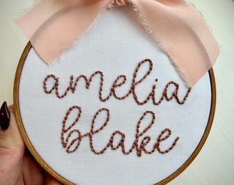 May include: A white embroidery hoop with a brown wooden frame. The hoop is decorated with a pink ribbon bow and features the embroidered text "amelia blake" in brown thread.