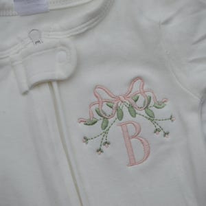 May include: Cream-colored baby sleeper with a pink embroidered bow, floral sprigs, and the letter "B". The sleeper has a full-length zipper and is made of soft fabric. A charming and personalized baby garment.