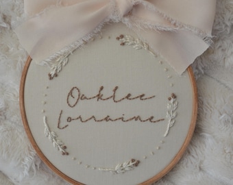 May include: A beige embroidery hoop with the name "Oaklee Lorraine" embroidered in brown thread. The hoop is decorated with small white and brown floral embroidery. A light pink ribbon is tied around the hoop.