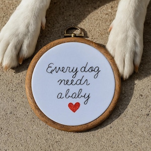 May include: A round embroidery hoop with the text "Every dog needs a baby" in black script, and a red heart. The hoop is made of wood and has a metal hanger. Two white dog paws are in the background.