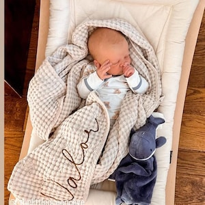 May include: A sleeping baby wrapped in a beige waffle-weave blanket with the name "Valor" embroidered on it. A blue plush whale toy rests beside the baby. The baby is lying on a cream-colored padded surface.