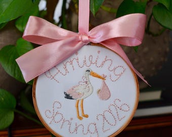 May include: Embroidered hoop art with a stork carrying a baby in a pink bundle. The text "Arriving Spring 2016" is stitched in pink thread. A pink ribbon is attached for hanging. The hoop is surrounded by green leaves.