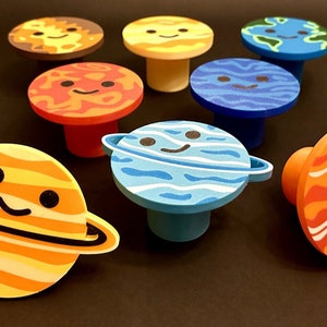 Op de afbeelding: Een verzameling grillige kastknoppen met een planetenthema. Elke knop heeft een lachend planeetontwerp, waaronder de zon, de aarde en Saturnus, in levendige kleuren. Deze decoratieve knoppen geven meubels een speelse uitstraling.