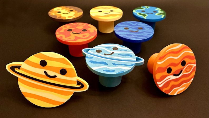 May include: A selection of planet-shaped cabinet knobs. Each knob depicts a smiling planet, such as the sun, Earth, and Saturn, in bright colours. These decorative knobs add a playful touch to furniture, measured in centimetres.