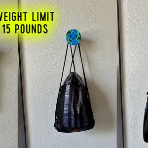 May include: Three wall hooks with a globe design, each holding a different item. The hooks are holding a blue and black dog leash, a black and grey drawstring bag, and a black and blue duffel bag. Text above reads "WEIGHT LIMIT 15 POUNDS."