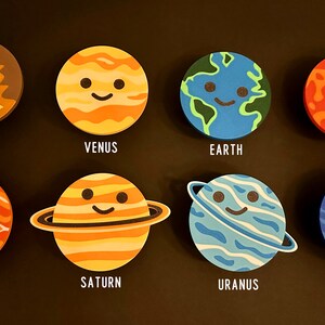 May include: Eight planet-shaped coasters with smiling faces and planet names: Mercury, Venus, Earth, Mars, Jupiter, Saturn, Uranus, and Neptune. The coasters are arranged on a black surface, ideal for space enthusiasts.