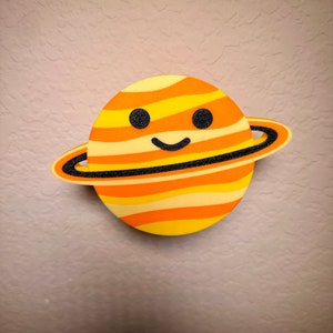 May include: A cartoon Saturn planet decoration with a smiling face. The planet is yellow, orange, and black striped, with a black ring. The decoration is likely made of foam or a similar material. A fun, decorative item for a child's room or space-themed decor.