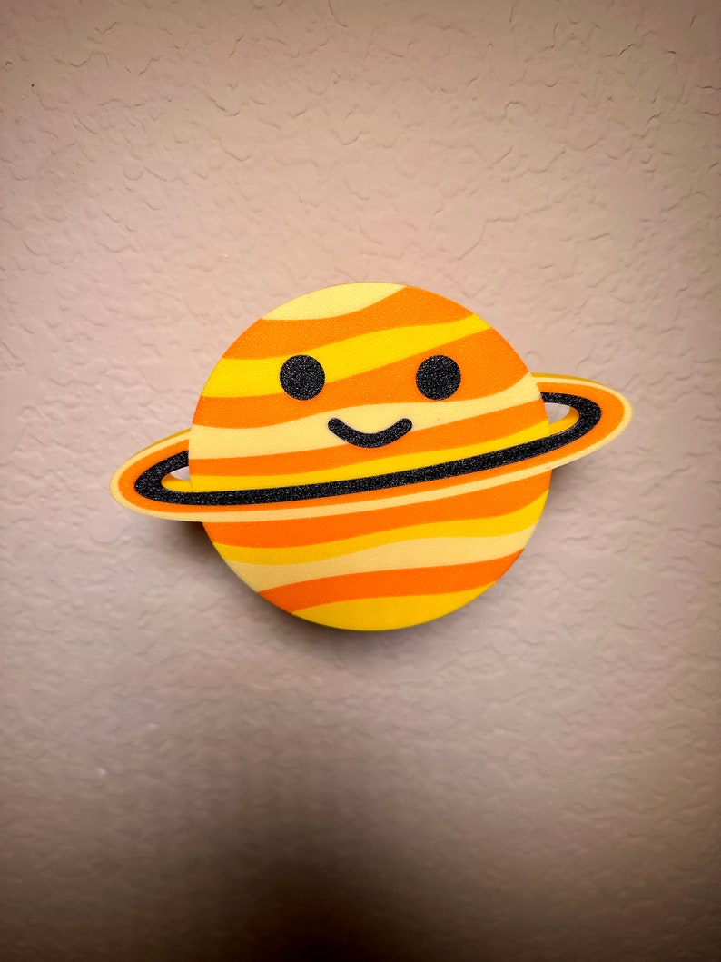 May include: A cartoon Saturn planet decoration with a smiling face. The planet is yellow, orange, and black striped, with a black ring. The decoration is likely made of foam or a similar material. A fun, decorative item for a child's room or space-themed decor.