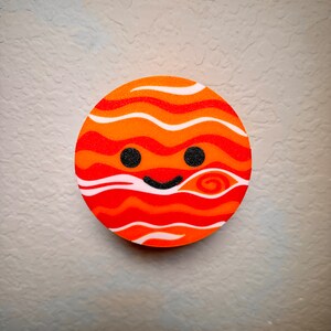May include: A circular design depicting a smiling planet in shades of orange and red. The planet features wavy stripes, two black dot eyes, and a curved black smile. The background is a textured, light grey surface, typical of a wall.