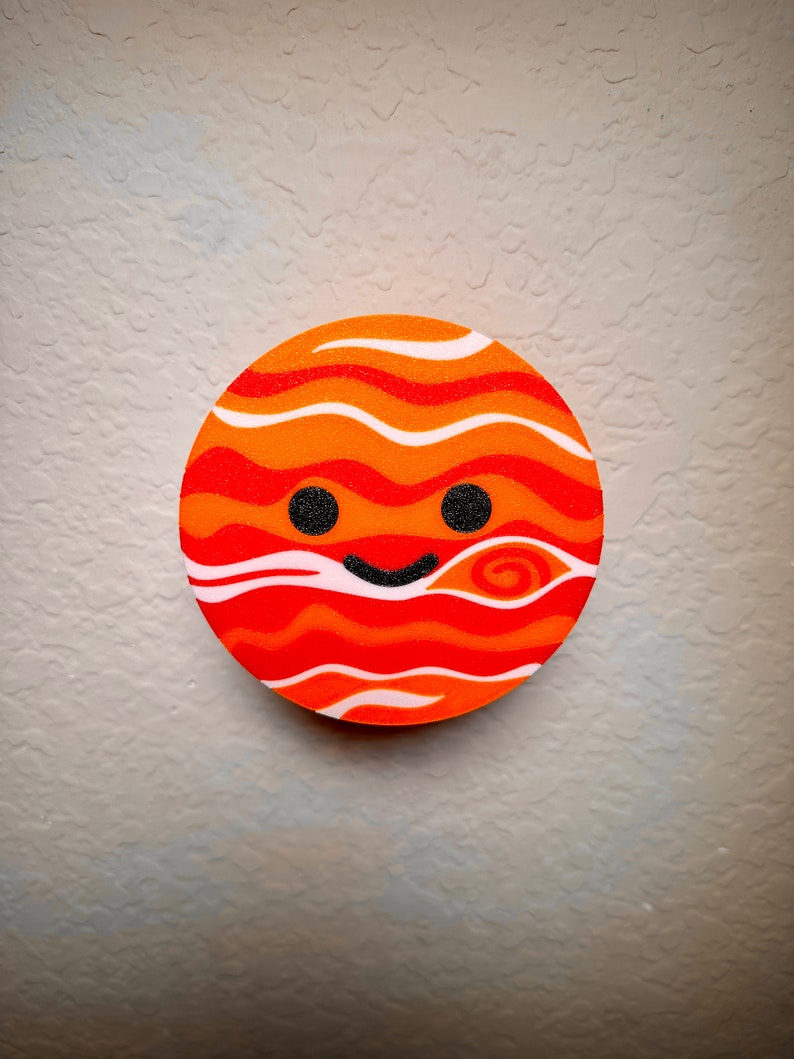 May include: A circular design depicting a smiling planet in shades of orange and red. The planet features wavy stripes, two black dot eyes, and a curved black smile. The background is a textured, light grey surface, typical of a wall.