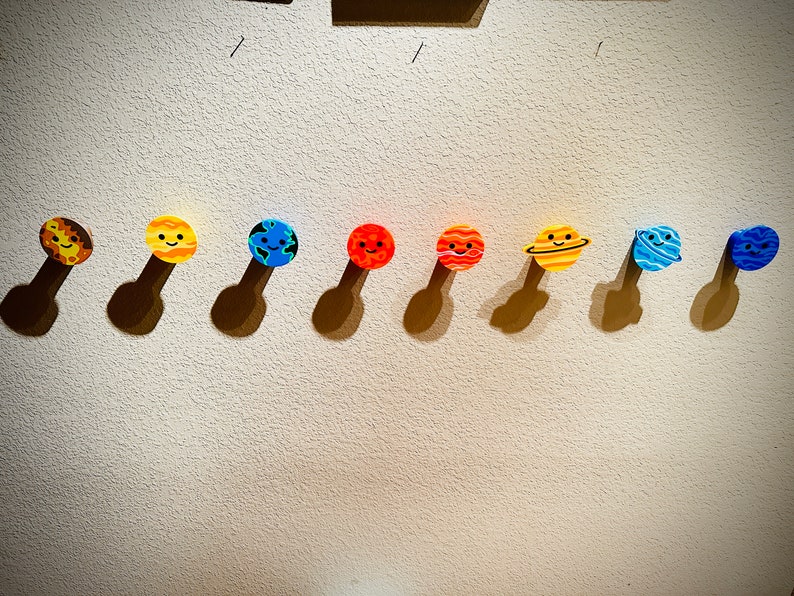 May include: Eight colourful planet decorations with smiling faces, each attached to a brown stem. The planets include Mercury, Venus, Earth, Mars, Jupiter, Saturn, Uranus, and Neptune. The decorations are mounted on a textured wall.