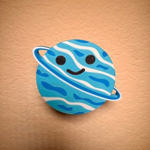May include: A blue planet-shaped decoration with a smiling face and a white ring. The planet features a wavy pattern in shades of blue and white, with black eyes. This whimsical item is designed for decorative purposes.