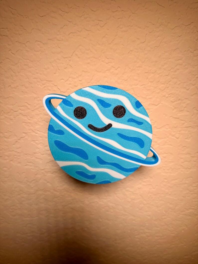 May include: A blue planet-shaped decoration with a smiling face and a white ring. The planet features a wavy pattern in shades of blue and white, with black eyes. This whimsical item is designed for decorative purposes.