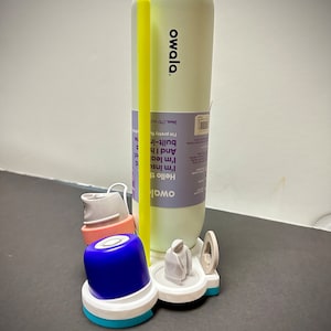 May include: A white and blue water bottle holder with a yellow straw, a blue lid, and a white lid. The holder has a white base with a blue ring around the edge. The bottle is white with the word "owala" printed on it in black. The text on the bottle reads "Hello there, I'm instantly leak-proof. And I have a built-in straw. I'm pretty much perfect. 24oz. (710 ml)"