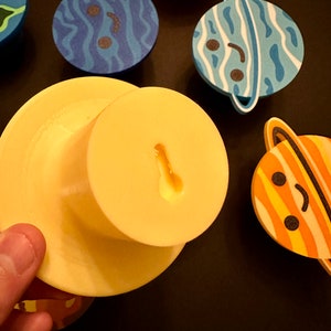 May include: A selection of planet-shaped magnets with smiling faces. The designs include blue and white stripes, orange and white stripes, and blue swirls. A yellow magnet with a keyhole shape is also visible. The magnets are for decorative purposes.