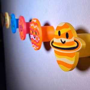 May include: A set of planet-themed wall hooks in a row. Each hook is a different colour and design, with smiling faces. The colours include orange, yellow, blue, and red. The hooks are mounted on a light-coloured wall.