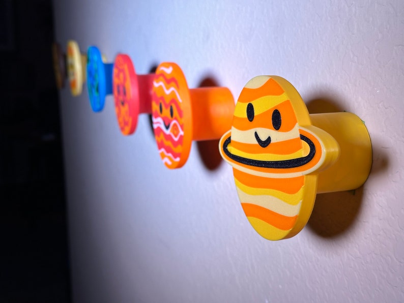 May include: A set of planet-themed wall hooks in a row. Each hook is a different colour and design, with smiling faces. The colours include orange, yellow, blue, and red. The hooks are mounted on a light-coloured wall.