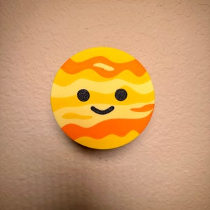 May include: A circular, yellow and orange planet-shaped decoration with a smiling face. The planet has a textured surface with orange stripes, two black dot eyes and a curved black mouth. The background is a textured beige wall.
