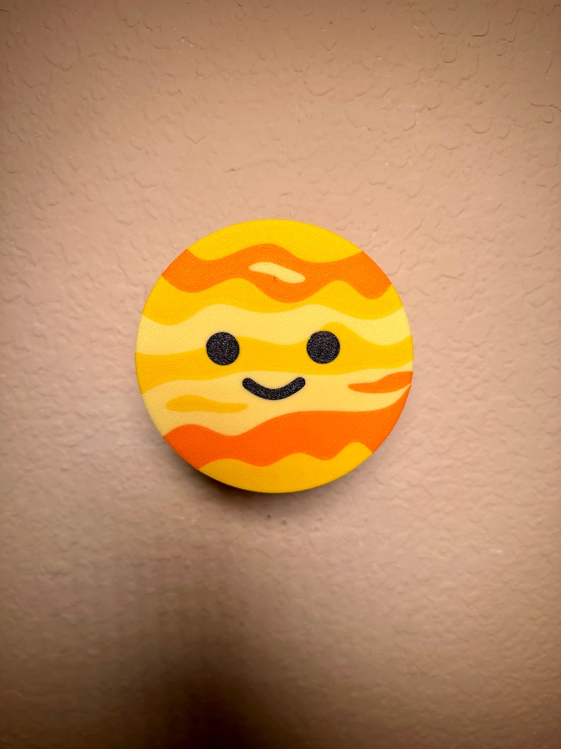 May include: A circular, yellow and orange planet-shaped decoration with a smiling face. The planet has a textured surface with orange stripes, two black dot eyes and a curved black mouth. The background is a textured beige wall.