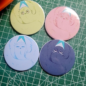 May include: Four round buttons with cartoon illustrations in pastel colors. Each button features a whimsical creature with a triangular hat. The buttons are in shades of yellow, pink, light blue, and dark blue, with a green cutting mat in the background.