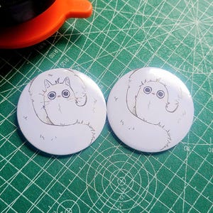 May include: Two white circular badges featuring a cartoon cat design. The cat has big eyes and a fluffy body. The badges are on a green cutting mat.