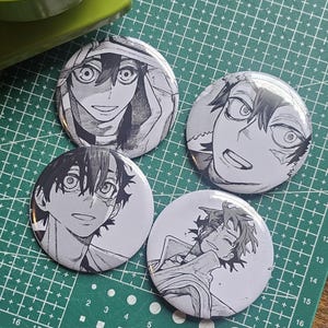 May include: Four round, black and white anime character buttons. Each button features a different manga-style illustration of a character's face. The buttons are on a green cutting mat with white grid lines.
