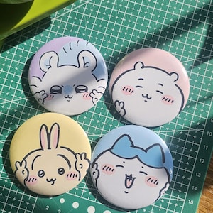 May include: Four cartoon character badges in pastel colours. Each badge features a different character with a unique expression and colour scheme: blue, pink, yellow, and light blue. The badges are on a green cutting mat.